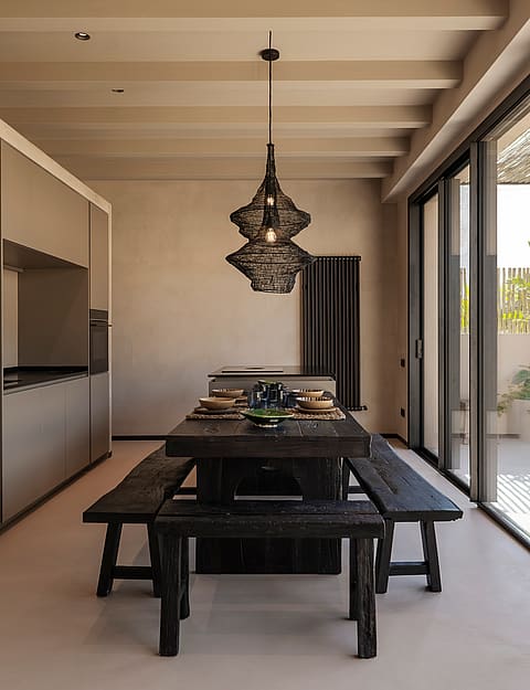 A modern kitchen and dining space featuring a dark, rustic wooden table with matching benches, a decorative pendant light, and sliding glass doors leading to an outdoor space.
