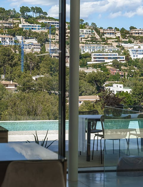 Interior view through floor-to-ceiling glass doors overlooking a terrace with a pool and the forested hills of Son Vida.