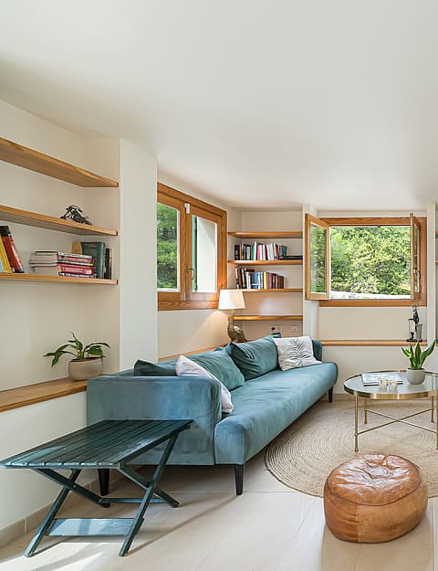 Bright living room featuring a blue velvet sofa, built-in wooden shelves, and casement windows showing garden greenery.