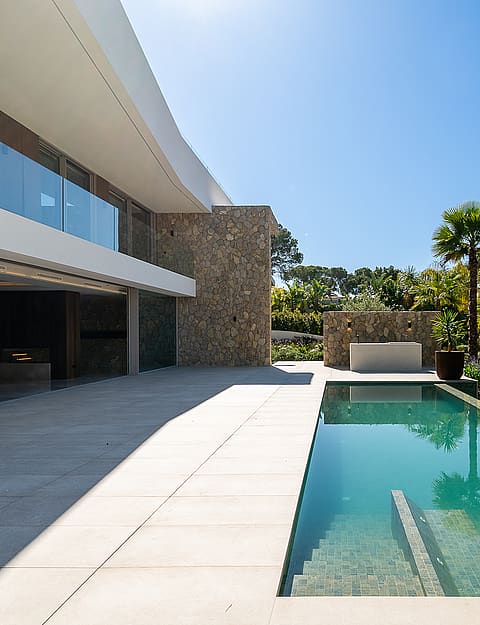 Modern villa exterior with a stone-clad wall, large terrace, and a rectangular swimming pool surrounded by palm trees.