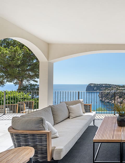 A covered outdoor lounge featuring gray sofas and wooden tables under white arches, overlooking the blue sea and cliffs.
