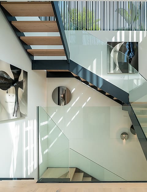 Modern open-tread staircase made of light wood and black metal, featuring frameless glass railings and vertical wooden slats.