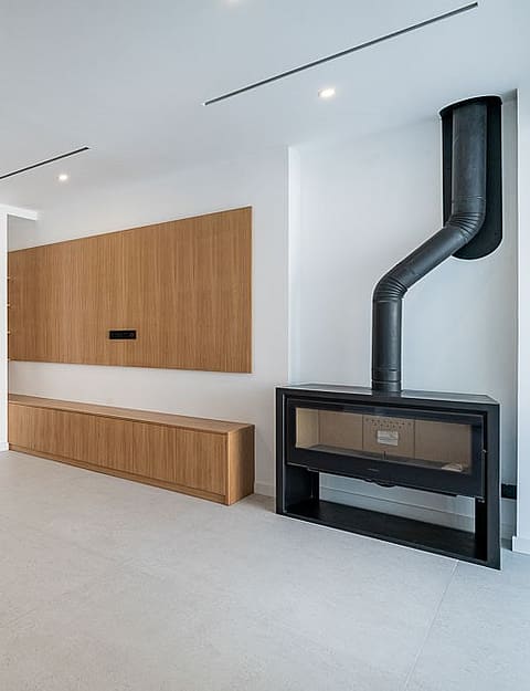 An expansive, bright living space with large light-grey floor tiles, custom light oak cabinetry, and a black metal fireplace.