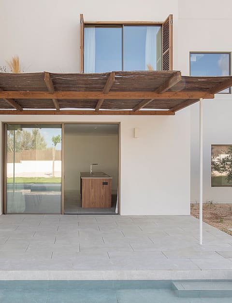 A stone terrace with a wooden pergola sits beside a swimming pool, leading to a modern white villa with glass doors.