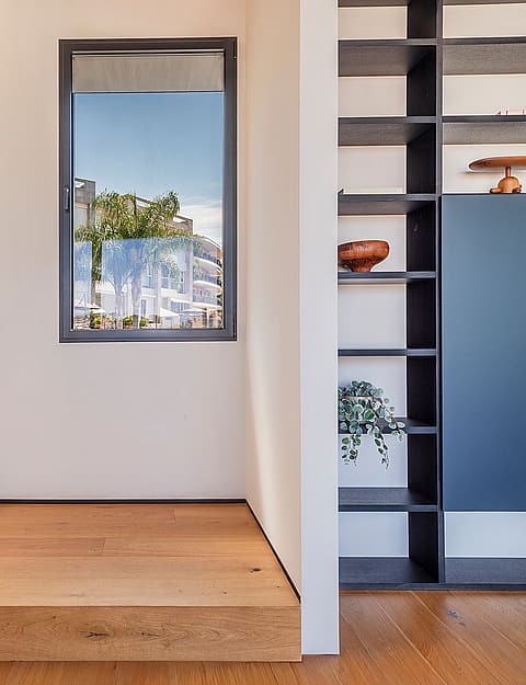 Interior wooden staircase with glass railing, light oak treads, and a built-in dark shelving unit beside a square window.