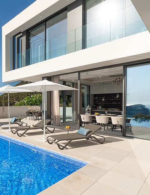 Modern villa with a blue tiled infinity pool and sun loungers on a stone terrace, set against a Mediterranean coastal landscape.