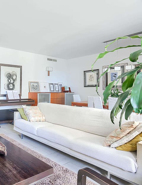 A bright living room with white walls, beige sofa, dark wood coffee table, and an armchair, leading to an interior hallway.