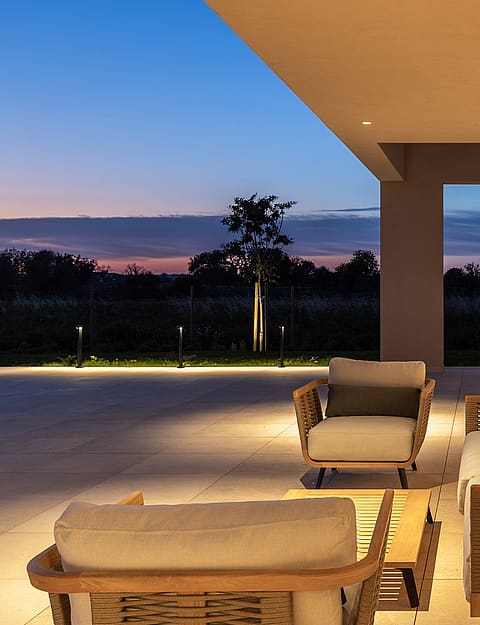 A covered stone terrace featuring woven outdoor seating with soft cushions during twilight, overlooking a dark garden.