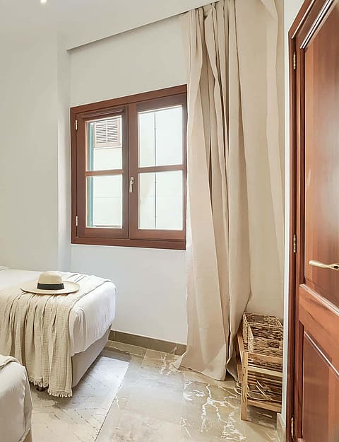 A bright bedroom with two twin beds, white walls, light tile flooring, and a wooden window frame with beige curtains.