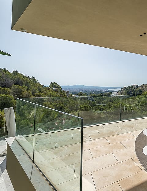 A wide, covered outdoor terrace overlooking hills and a distant coastline, featuring neutral outdoor furniture, glass railings, and stone flooring.