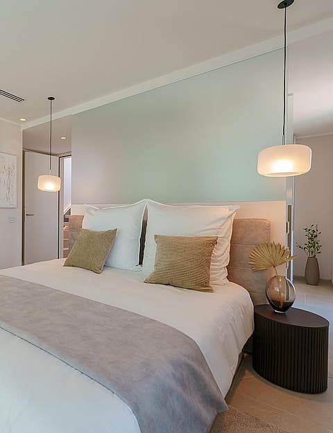 Dormitorio luminoso con cama de matrimonio, ropa de cama blanca, cojines neutros e iluminación colgante.