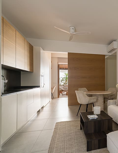 A bright apartment living space featuring a white sofa, minimalist kitchenette with wood cabinets, and dining area.
