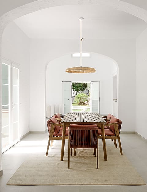 Comedor con bóveda blanca, mesa de teca, sillas de cuerda y lámpara de ratán, vistas al jardín a través de puertas abiertas