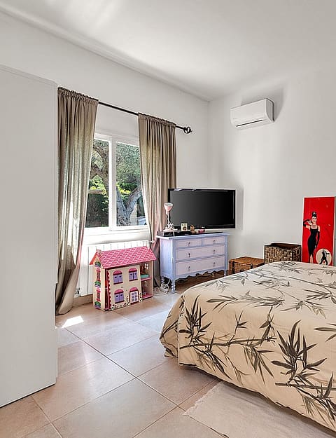 A bedroom with white walls, a patterned beige quilt on the bed, a large carved wooden headboard, and a window with curtains.