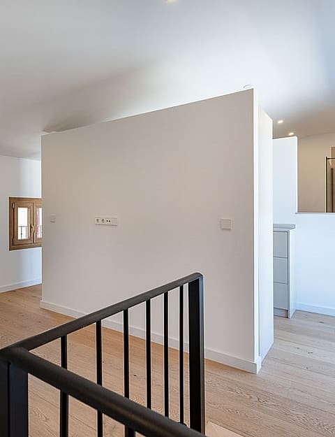 An open landing area in a renovated townhouse with white walls, light wood flooring, a black metal staircase railing, and wooden window frames.