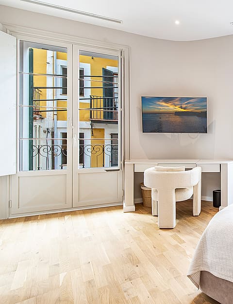 Bright bedroom with white walls, light wood floors, a desk, and floor-to-ceiling windows looking out onto a yellow building.