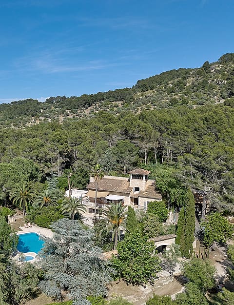 Aerial shot of a stone finca surrounded by dense green forest and mountains under a clear blue sky, with a visible pool.
