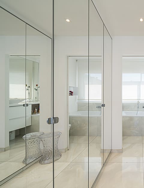 A bright dressing room featuring wall-to-wall mirrored wardrobes with chrome handles, leading into a white tiled bathroom.