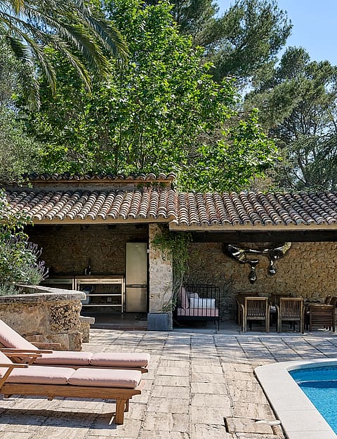 A stone-paved patio with two wooden sun loungers beside a swimming pool, featuring a covered outdoor dining area with a stone wall and rustic tiled roof.