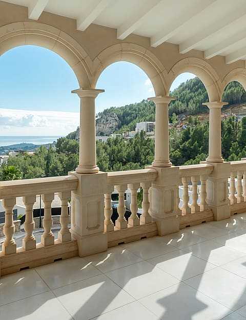 A covered stone loggia with arched columns and a balustrade overlooking a green valley and distant coastal city under clear skies.