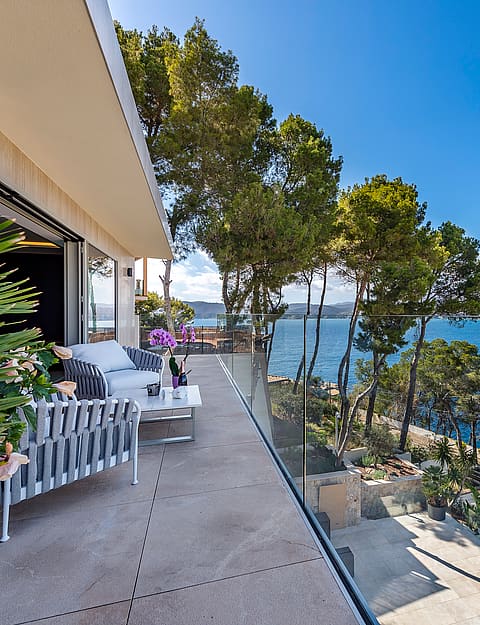 A wide terrace with grey outdoor furniture, glass railings, and a view overlooking the Mediterranean coastline and pines.