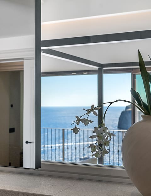 A clay vase with orchids reflects a metal bed frame and the blue sea.