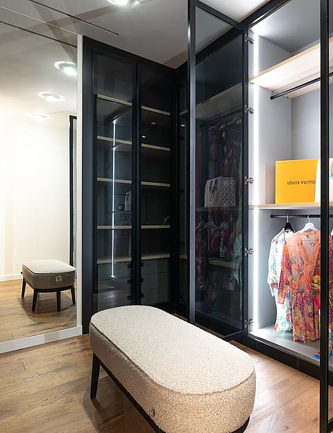 Walk-in closet with dark metal and glass cabinetry, integrated LED lighting, and a textured ottoman on hardwood flooring.