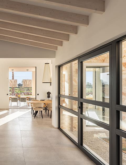 Interior hallway of a stone finca with large black-framed glass doors, light stone floors, and high wood-beamed ceilings.