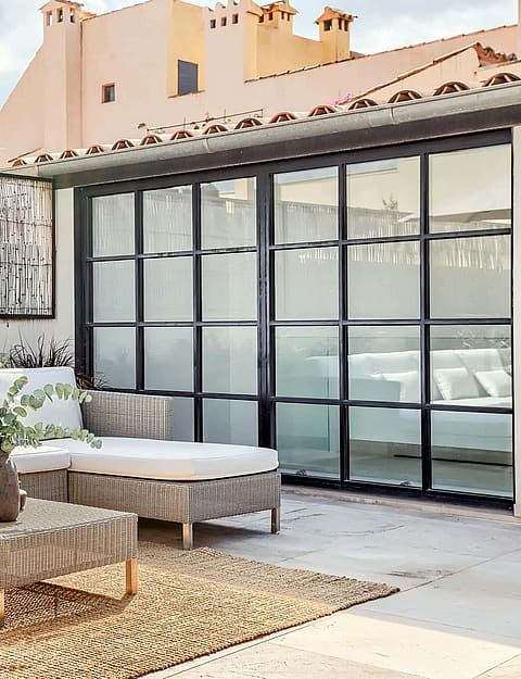 A private outdoor terrace with woven beige sofa sets, a coffee table on a natural fiber rug, and black steel-framed glass doors.