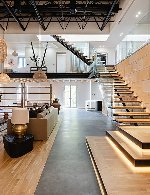 Double-height living room featuring a floating wooden staircase, limestone feature wall, and industrial black steel ceiling trusses.