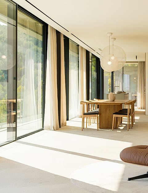 A bright living space featuring a brown leather lounge chair, wooden dining table, and large glass walls looking outside.