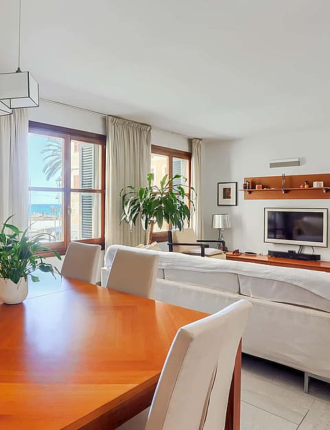 A bright living and dining room with wooden furniture, white sofas, and windows overlooking the sea.