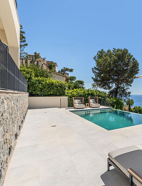 Infinity pool on a light stone terrace with sun loungers overlooking the blue sea under a clear sky.