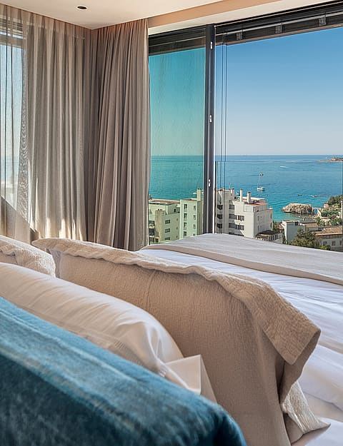 A view from inside a bedroom looking through floor-to-ceiling glass sliding doors at a Mediterranean coastline and sea.