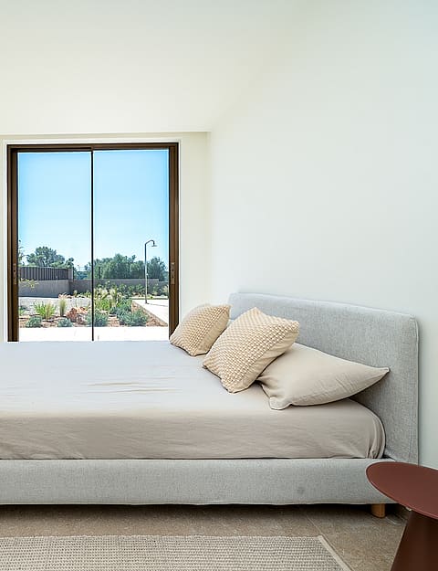 A minimalist bedroom featuring a grey upholstered bed, floor-to-ceiling white wardrobe, and a sliding glass door to a garden.
