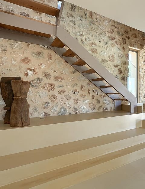 A modern staircase with wooden treads ascends against a feature wall made of exposed natural stone in a bright finca interior.