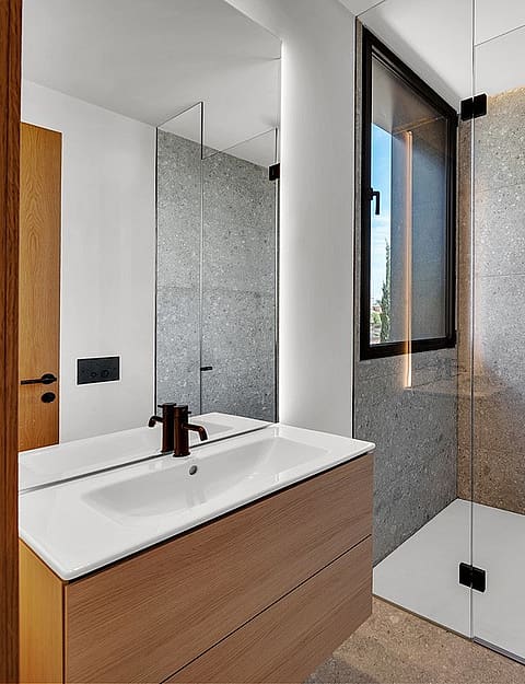 Interior view of a modern bathroom featuring a white vanity with wooden cabinetry, a large wall mirror, and a walk-in shower with grey stone tiles and black fixtures.