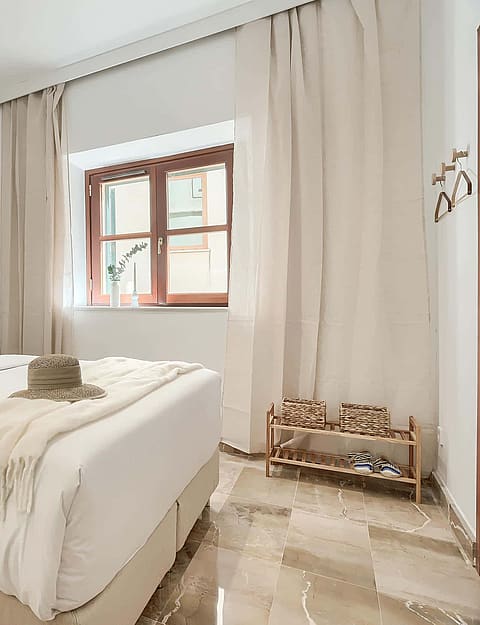 A bright bedroom with a queen-sized bed, white linens, beige curtains, and polished marble flooring in an Old Town townhouse.