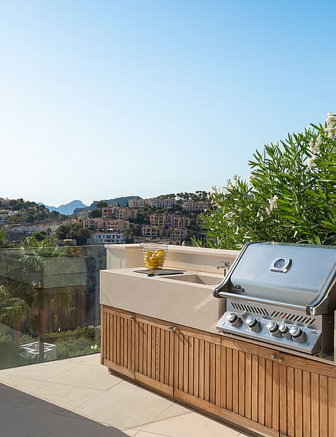 A stainless steel barbecue grill integrated into a light-toned stone and wood kitchen unit on a sunny residential terrace.
