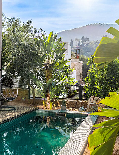 Outdoor swimming pool with turquoise water and pebble deck adjacent to a beige villa with lush green garden and mountain views.