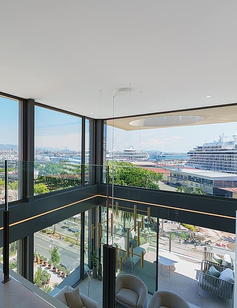 Salón de doble altura con paredes de cristal, una butaca de terciopelo y vistas al puerto.
