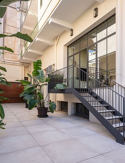 A private outdoor courtyard with light stone paving, lush potted plants, and a black metal staircase leading to a glazed entrance.