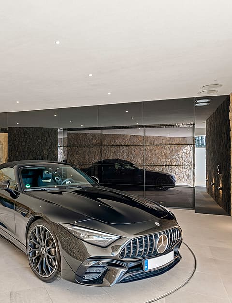 A luxury garage interior featuring a dark sports car on a mechanical turntable, flanked by natural stone walls and planters.