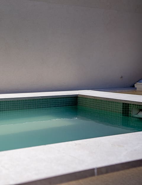 A rectangular plunge pool featuring green mosaic tiles and light-colored stone coping against a minimalist white wall.