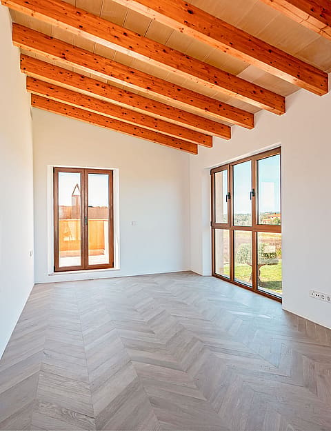 An empty, bright bedroom with white walls, herringbone parquet flooring, and exposed wooden ceiling beams, featuring large windows.