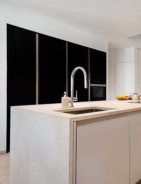 Modern kitchen with a light stone island, matte black cabinetry, and a floating staircase with thin metal railings.