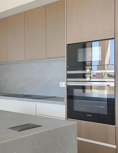 A modern kitchen with a grey stone-topped island, light wood cabinetry, and a double wall oven next to a window.