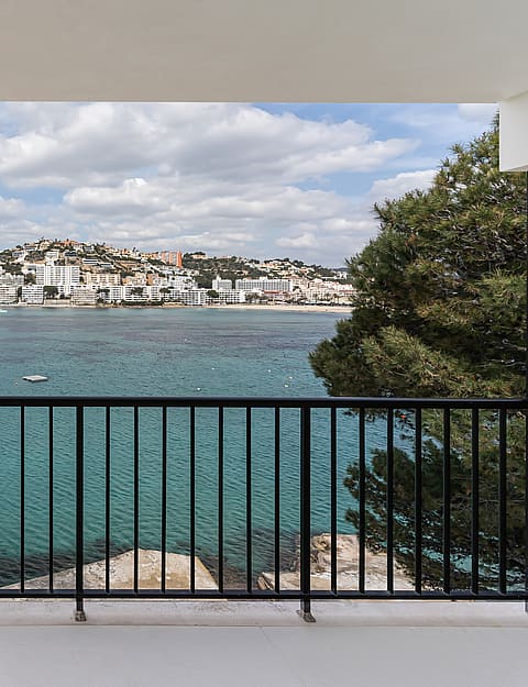 A balcony with black metal railing overlooking a turquoise bay and white coastal town under a soft sky.
