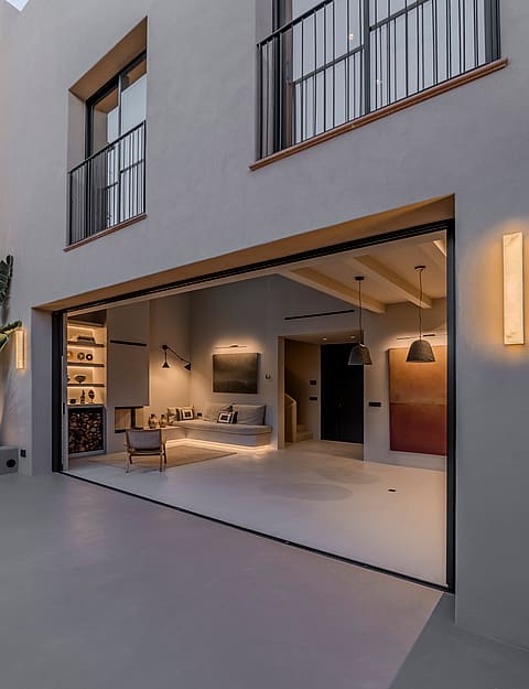 Exterior shot of a white townhouse facade with wide sliding doors revealing an interior with wood beams and a fireplace.