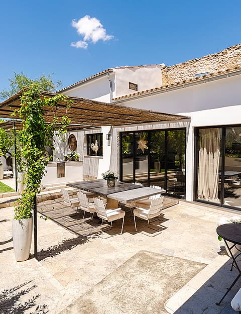 A sunny stone-paved terrace with a wooden pergola covering a dining table and chairs, adjacent to a white house facade.